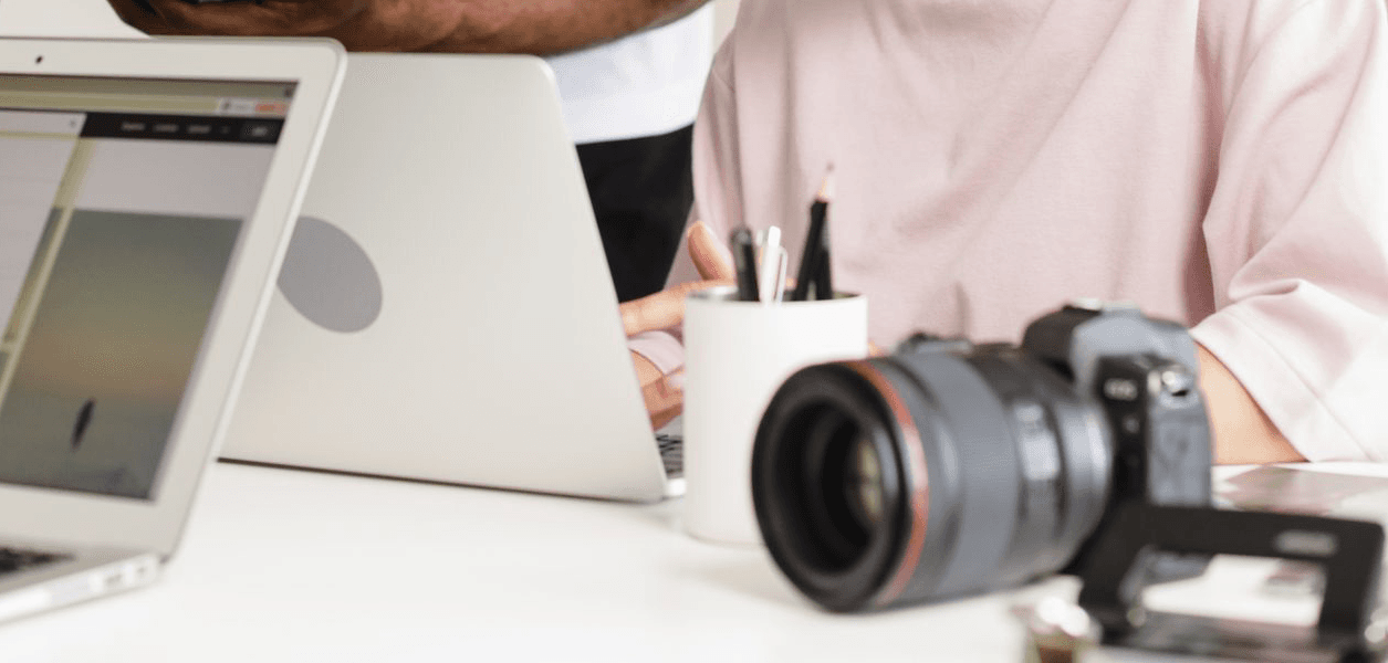 Two people working at a desk with a laptop, a camera and a pot of pencils in the foreground