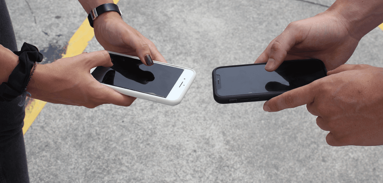 Two people standing outdoors holding out their mobile phones toward each other