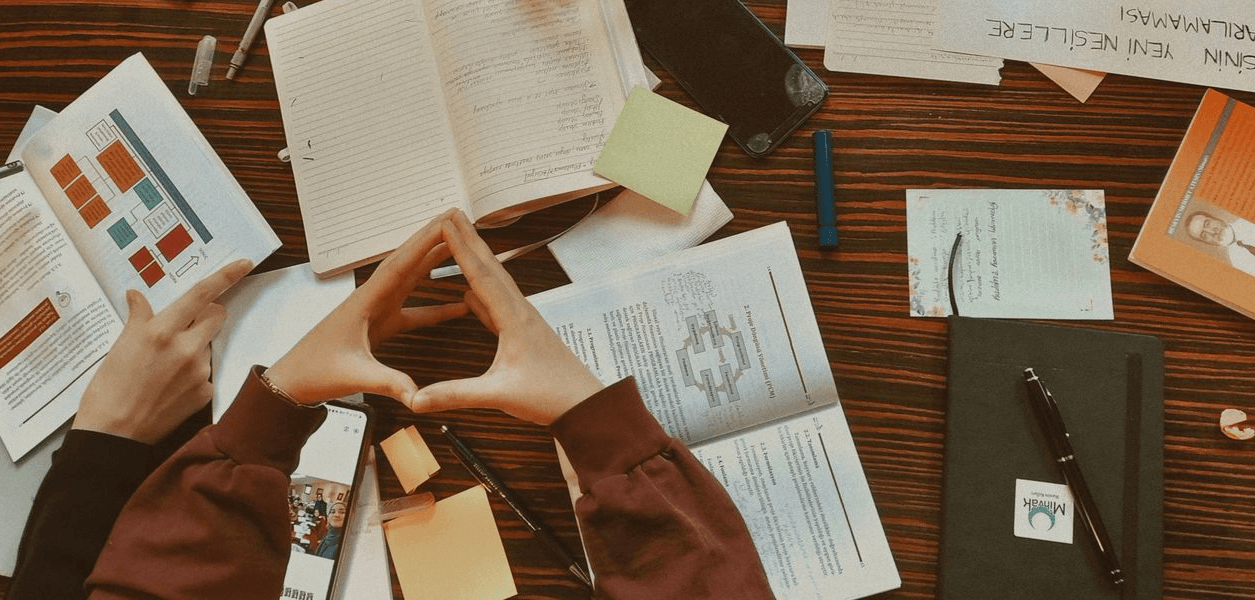 Study desk seen from above with open notebooks, printed research and sticky notes; a pair of hands forms a triangle over the pages