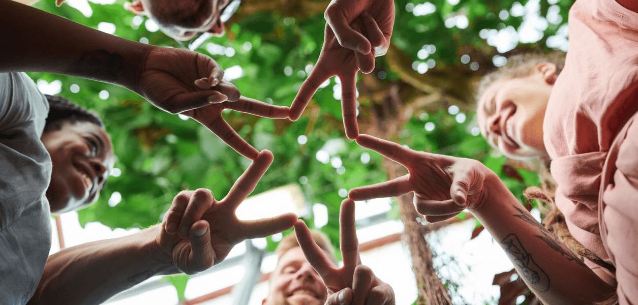 Five people photographed from below, touching fingertips together to form a star under a sunlit tree